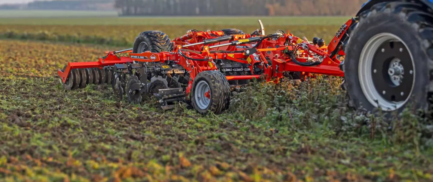 Kuhn's stubble disc cultivator Optimer XL 5000 being pulled by a tractor across a ploughed field, with rolling hills and forests visible in the background under an overcast sky.